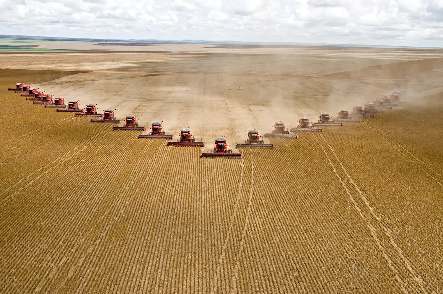 Aerial photograph of 25 combine harvesters driving in V formation across a soybean field.